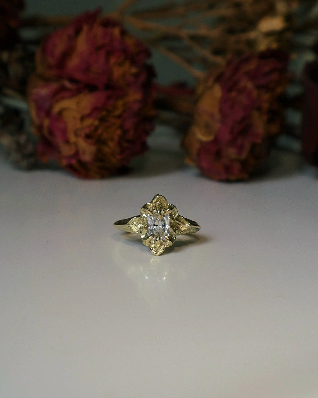 Gold ring with a diamond on a white surface with dried flowers in the background